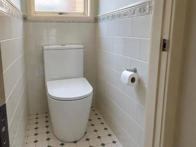 White toilet in a small tiled bathroom with patterned border and frosted window in Lambton